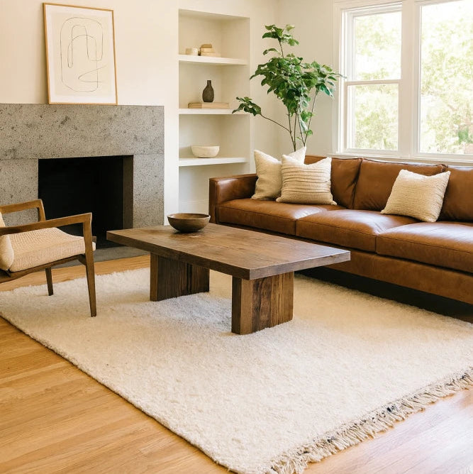 Le Tapis Berbère Uni Béni Ouarain de MAZIR Décoration donne du charme à un salon moderne composé d'un canapé en cuir marron, d'une table basse en bois, d'un tapis crème et d'œuvres d'art abstraites.