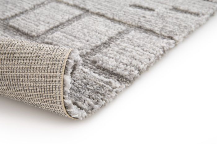 Tapis moderne berbère gris avec motif géométrique en relief, texture moelleuse et dos tissé visible, partiellement enroulé.