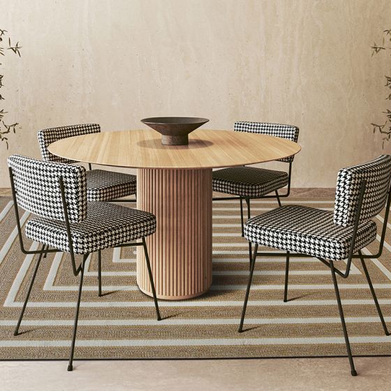 Tapis en jute à motif géométrique de rayures beiges et blanches, placé sous une table ronde en bois et des chaises à motif pied-de-poule.