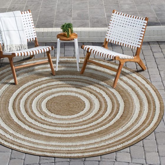 Tapis rond en jute naturel beige avec des cercles concentriques crème, posé sur un patio en pavés gris avec des chaises et une table d'appoint.